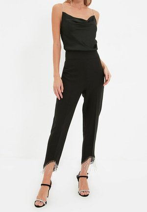 Mujer con top negro sin mangas con tirantes de cadena y pantalones negros con flecos de cuentas en el dobladillo, posando con las manos relajadas.