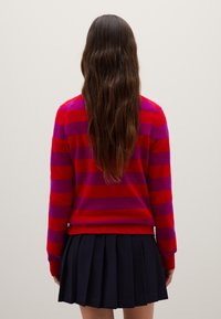 Roter und lila gestreifter Pullover mit langen Ärmeln und rundem Halsausschnitt, kombiniert mit einem dunklen Faltenrock. Weiches Material und lockere Passform.