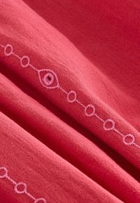 Tissu corail avec une finition texturée, présentant des cercles brodés rose pâle et des motifs en losange le long d'une ligne courbe.