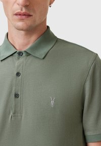 Polo in verde oliva, realizzato in un tessuto testurizzato. Presenta un colletto con bottoni e un piccolo teschio di capra ricamato sul petto.