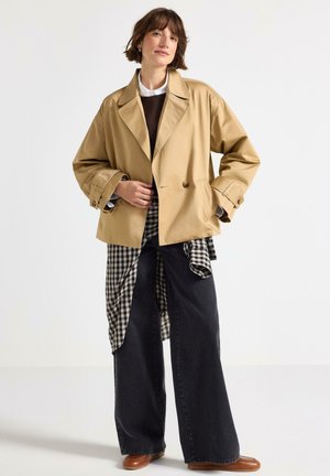 Giacca beige oversize con design doppiopetto, abbinata a un maglione marrone e a una camicia nera a quadri, indossata su jeans neri larghi.