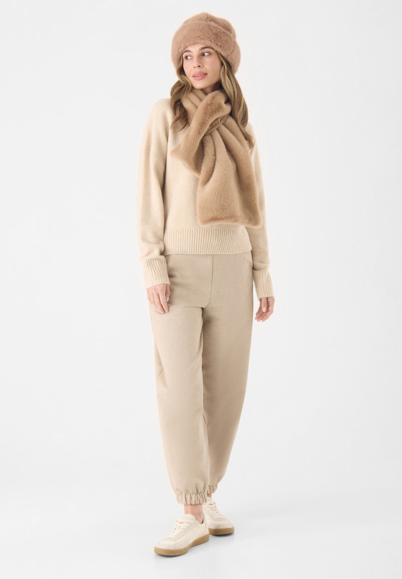 Beige trui en bijpassende joggingbroek, gecombineerd met een zachte beige sjaal en muts. De stof lijkt zacht en heeft een relaxte pasvorm met geribbelde manchetten.