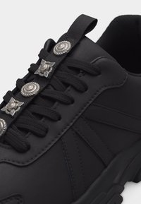 Μαύρο αθλητικό παπούτσι με παχιά σόλα, μαύρα κορδόνια και ασημένια διακοσμητικά μεταλλικά κουμπιά κατά μήκος του λουριού της γλώσσας.