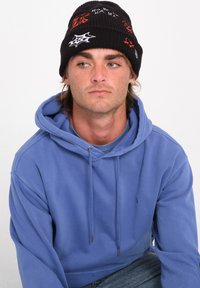 Berretto nero con design ricamati in bianco e rosso, inclusi ragnatele. Indossato con una felpa leggera blu. Aspetto semplice e casual.