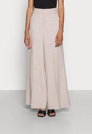 Kobieta w beżowych, szerokich, wysokich spodniach, czarnym topie i czarnych, otwartych na palce szpilkach, stojąca na jasnym, jednolitym tle.