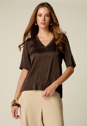 Frau mit langen braunen Haaren trägt ein dunkelbraunes Satin-V-Ausschnitt-Top, beige Hose mit Spitzen-Details, goldene Ohrringe und breite Armbänder am linken Handgelenk.