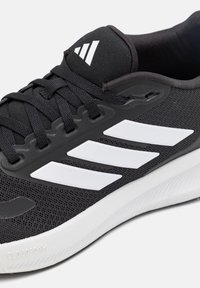 Chaussure de course Adidas noire avec des bandes blanches et une semelle blanche avec un amorti Cloudfoam, gros plan sur les lacets et le panneau latéral.