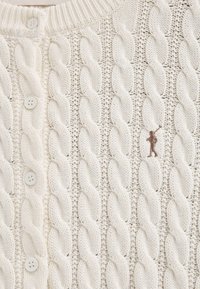 Weiße Strickjacke mit Kabelmuster, mit geripptem Ausschnitt, fünf Knöpfen vorne und einer kleinen braunen Stickerei.