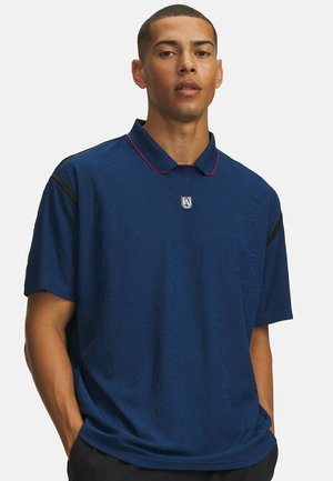 Man met kort haar draagt een donkerblauw poloshirt met subtiele patronen, een klein logo op de borst en de handen in de zakken.