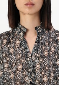 Μαύρη διάφανη μπλούζα με διακοσμητικό μοτίβο σε μπεζ, γκρι και σκουριά. Διαθέτει ριχτό γιακά και κλείνει με κουμπιά στο μπροστινό μέρος.