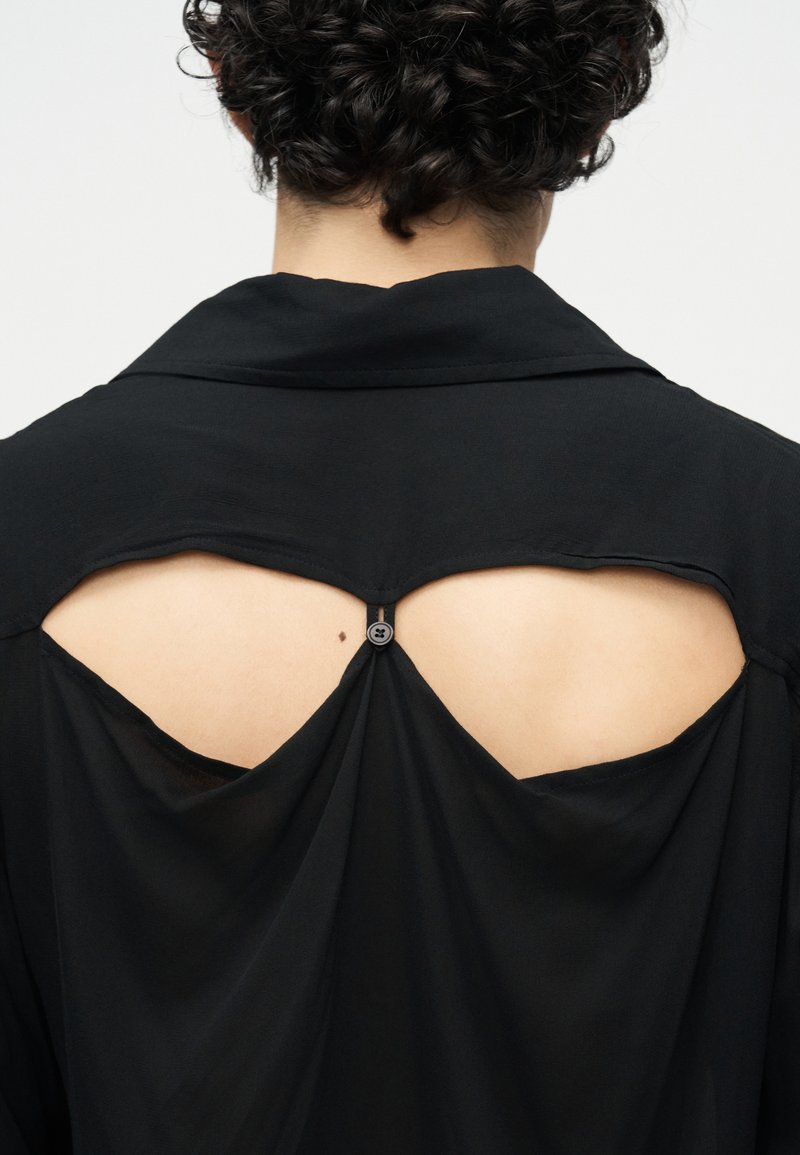 Person with short curly black hair wears black top featuring back cutouts fastened with a small button near the neck.