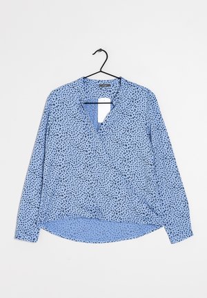 Lichtblauwe blouse met lange mouwen met zwart gespikkeld patroon, V-hals en afgeronde zoom, hangend aan een zwarte hanger tegen een witte achtergrond.