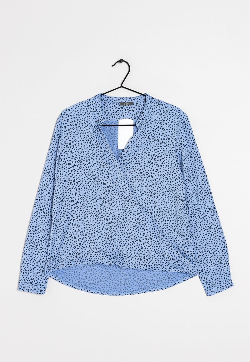 Blouse bleu clair à manches longues avec motif noir tacheté, col en V et ourlet arrondi suspendue sur un cintre noir sur fond blanc.