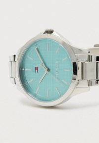 Sudraba metāla rokas pulkstenis ar gaiši zilu seju, sudraba stundu rādītājiem, trim rokām un Tommy Hilfiger logotipu uz baltā fona.