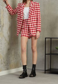 Röd och vit houndstooth-mönstrad blazer med matchande shorts. Har guldknappar och bärs med svarta ankelstövlar. Texturerad tyg.