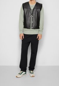 Svart läderväst över en ljusgrön sweatshirt, kombinerat med svarta byxor och vita sneakers med gröna detaljer. Enkelt mönster och avslappnad passform.