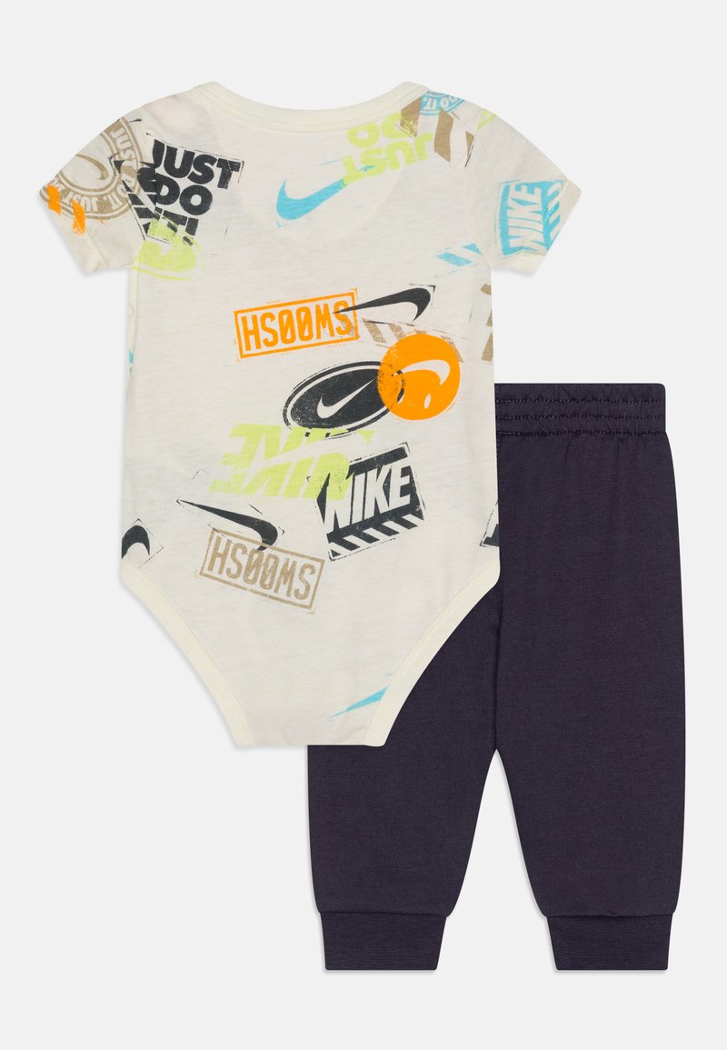 Ubranie dla dzieci składające się z kremowego body z krótkim rękawem z wielokolorowymi nadrukami logo oraz granatowych spodni z elastycznym paskiem i mankietami.