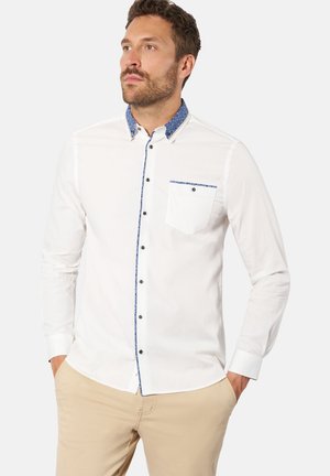 Mann trägt ein weißes Langarmhemd mit blauem Besatz am Kragen, an der Knopfleiste und an der Tasche, kombiniert mit beigen Hosen, Hände in den Taschen.