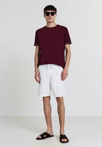 Uomo con occhiali da sole scuri, maglietta bordeaux a maniche corte, pantaloni corti bianchi con cordino e sandali intrecciati neri in piedi davanti a uno sfondo neutro.