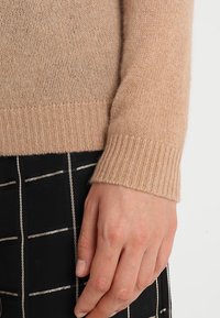 Beżowy kaszmirowy sweter z prążkowanym mankietem, ukazujący drobny splot dzianiny. Ręka położona na czarnej tkaninie z białym wzorem w kratkę.