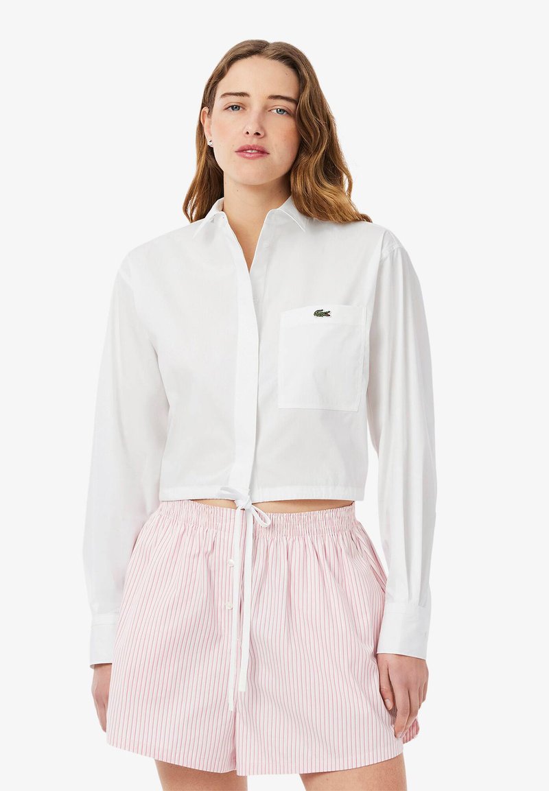 Camicia bianca corta con bottoni, maniche lunghe e taschino sul petto. Abbinata a pantaloni corti elasticizzati a vita alta a righe rosa e bianche.