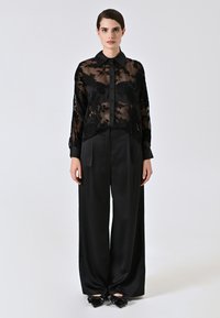 Zwarte sheer blouse met bloemenborduurwerk, knoopsluiting en lange mouwen, gecombineerd met hoge zwarte satijnen broek en schoenen met strikaccent.