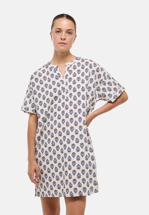 Femme aux cheveux attachés portant une robe blanche à manches courtes, longueur au genou, avec un motif floral bleu et beige sur fond blanc.