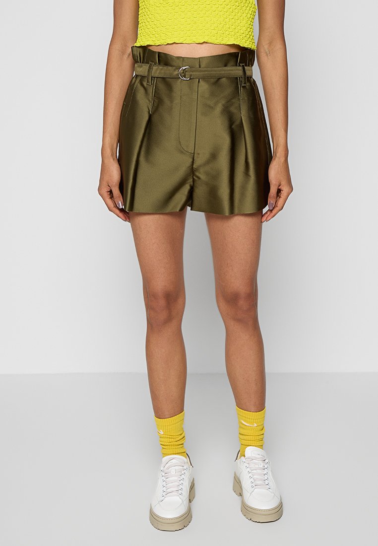 Olijfgroene satijnen shorts met een riem in de taille, voorplooien en zijzakken, gecombineerd met een felgeel cropped top en witte sneakers.