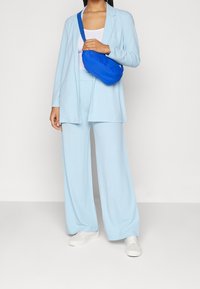 Lichtblauwe kostuum in zachte stof, met een relaxte blazer en wijde broekspijpen. Gepaarde met een blauwe crossbody tas en witte sneakers.