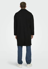 El abrigo de lana negro presenta un diseño entallado con un cuello, que llega a la mitad del muslo. Se combina con pantalones de rayas azul claro y zapatillas blancas.