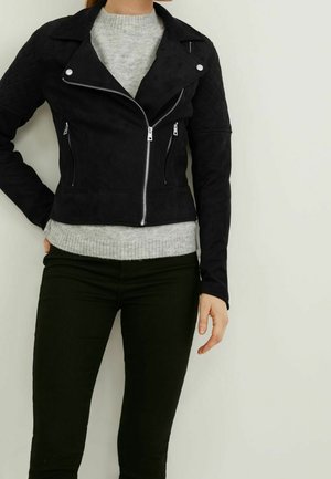 Person iført en sort bikerjakke med lynlås og quiltede ærmer over en grå sweater og sorte bukser, stående op ad en ensfarvet lys væg.