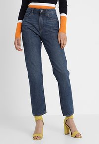 Vaqueros de mezclilla azules de cintura alta con corte recto. Combinados con sandalias de tacón en un vibrante color amarillo y un suéter negro con rayas naranjas.