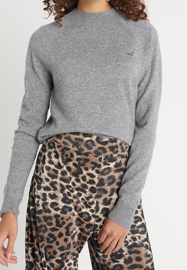 Femme portant un pull gris à manches longues et un pantalon taille haute à imprimé léopard, debout devant un fond clair uni.