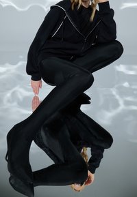 Felpa nera con dettagli bianchi sulla cerniera, abbinata a leggings neri e tacchi a punta. Il modello è seduto su una superficie riflettente, creando un effetto visivo sorprendente.