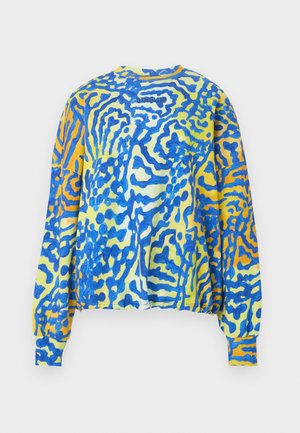 Vrouw met lang bruin haar, die een loszittende, kleurrijke blauw-gele trui met patroon, wijd uitlopende blauwe jeans en zwarte schoenen draagt, staand met haar handen achter haar rug.