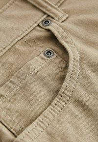 Tissu beige à texture lisse, avec une poche incurvée et des fermetures à boutons ornées d'accents métalliques et des détails de coutures visibles.