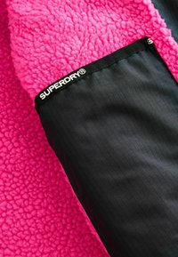 Újrahasznosított anyagból készült rózsaszín polárdzseki, fekete sima anyag részlettel az ujjon és a "Superdry" logóval.