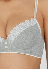 Šedý bralet s krajkovým lemem kolem výstřihu, se dvěma dekorativními knoflíky vpředu a tenkými ramínky. Měkká textilie.
