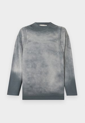 Mand iført grå langærmet skjorte, løse sorte cargobukser med store lommer, sorte sko og solbriller, stående med hænderne i lommerne.