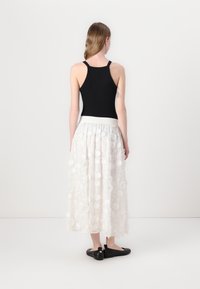 Zwarte mouwloze top gecombineerd met een witte maxi skirt met bloemapplicaties, elastische tailleband en zwarte platte schoenen.