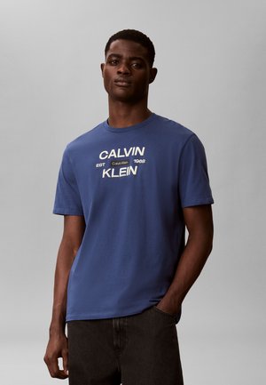 Homme portant un t-shirt Calvin Klein bleu avec texte du logo blanc, debout devant un fond gris uni, une main dans la poche.