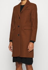 Brun uldblandet frakke med spidse klapper, fire knapper, to forlommer og en glat tekstur, lagdelt over en sort top og nederdel.