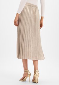 Femme portant une jupe midi plissée beige et des sandales à talons hauts métalliques dorées, debout devant un fond blanc.