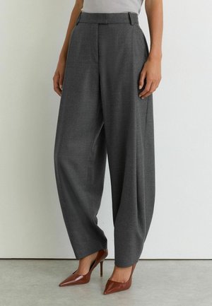 Person trägt hoch taillierte, weit geschnittene dunkelgraue Hose und braune spitze High-Heels und steht vor einer schlichten weißen Wand.