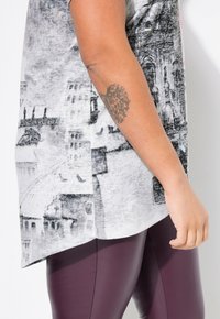 Camiseta gris de manga corta con un patrón de paisaje urbano, combinada con leggings morados oscuros. La camiseta tiene un dobladillo curvado y una textura suave.