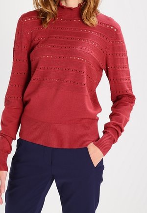 Femme portant un pull en maille rouge avec des rayures à motifs et des manches longues, associé à un pantalon bleu marine foncé, main dans la poche.