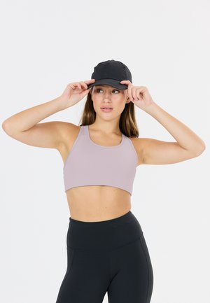 Junge Frau passt schwarze Mütze an, trägt ein hellviolettes Sport-BH und hoch taillierte schwarze Leggings, steht vor einem schlichten Hintergrund.