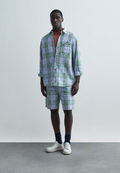 Ένδυμα με πολύχρωμο καρό σχέδιο που περιλαμβάνει πουκάμισο με κουμπιά και ταιριαστά σορτς, κατασκευασμένα από ελαφρύ ύφασμα. Περιλαμβάνει μπλε λεπτομέρειες και λευκά αθλητικά παπούτσια.