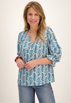 Vrouw met lang blond haar, gouden creolen, een blauw gebloemde blouse en lichtblauwe broek, glimlachend met de handen in elkaar geslagen.