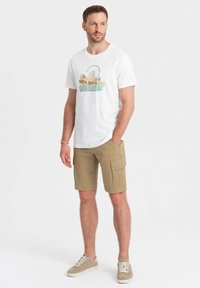 Maglietta bianca in cotone con una grafica colorata e testo, abbinata a pantaloni cargo beige con tasche laterali e taglio dritto. Sneakers casual.